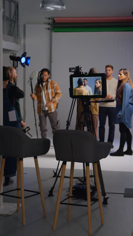 Vertical-Video-Of-Female-Director-Walking-Onto-Busy-Film-Set-With-Crew-Shooting-Movie-In-Studio-To-Talk-With-Actors